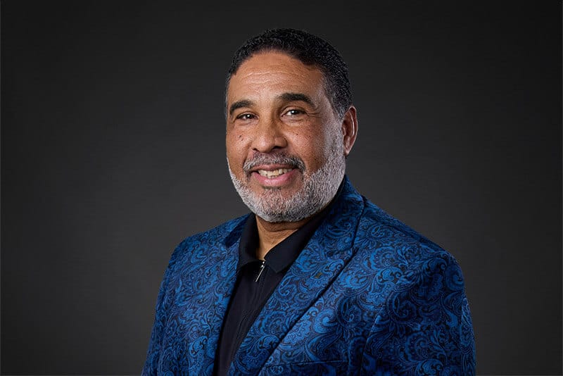 Professional headshot of a smiling man in a blue patterned suit, dark background, Project Big Apple partner.