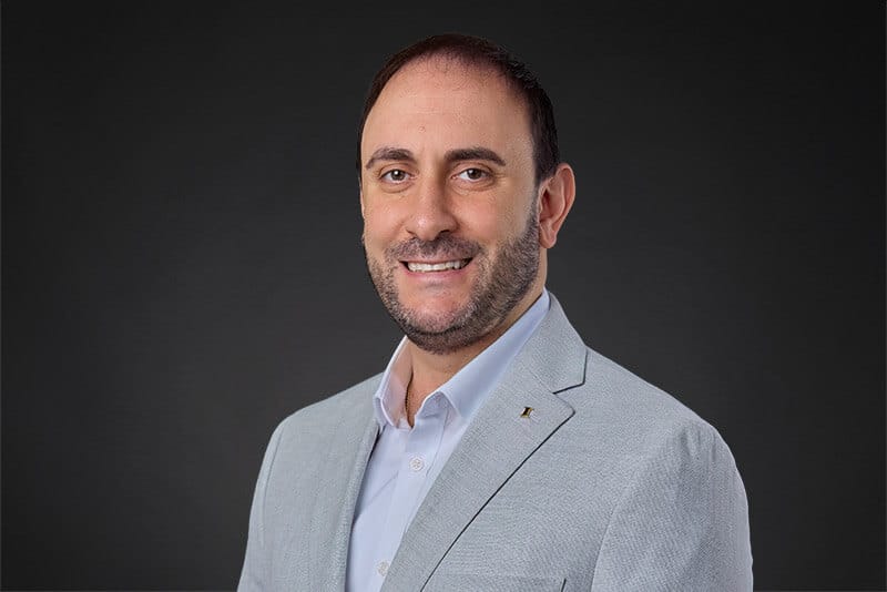 Professional headshot of a smiling man in a light gray suit, dark background, Project Big Apple partner portrait.
