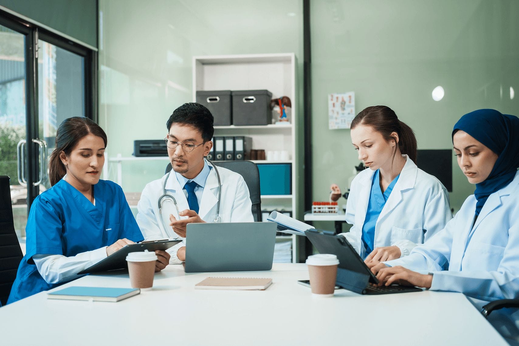 Diverse group of healthcare professionals in discussion, working on laptops and tablets in a modern medical office.